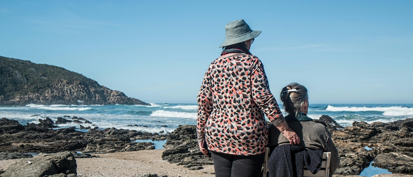 Two women on a beach. One is standing the other sits in a wheelchair.